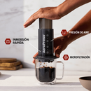 Manos presionando una cafetera AeroPress sobre una taza de vidrio, con etiquetas de inmersión, presión y microfiltración.