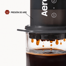Detalle de una AeroPress con gotas de café cayendo en una taza de vidrio y una etiqueta de presión de aire.