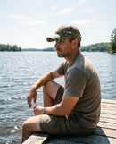 Hombre en el muelle de un lago bajo el sol con la gorra de camuflaje Ridgeline y el ala que le protege la cara